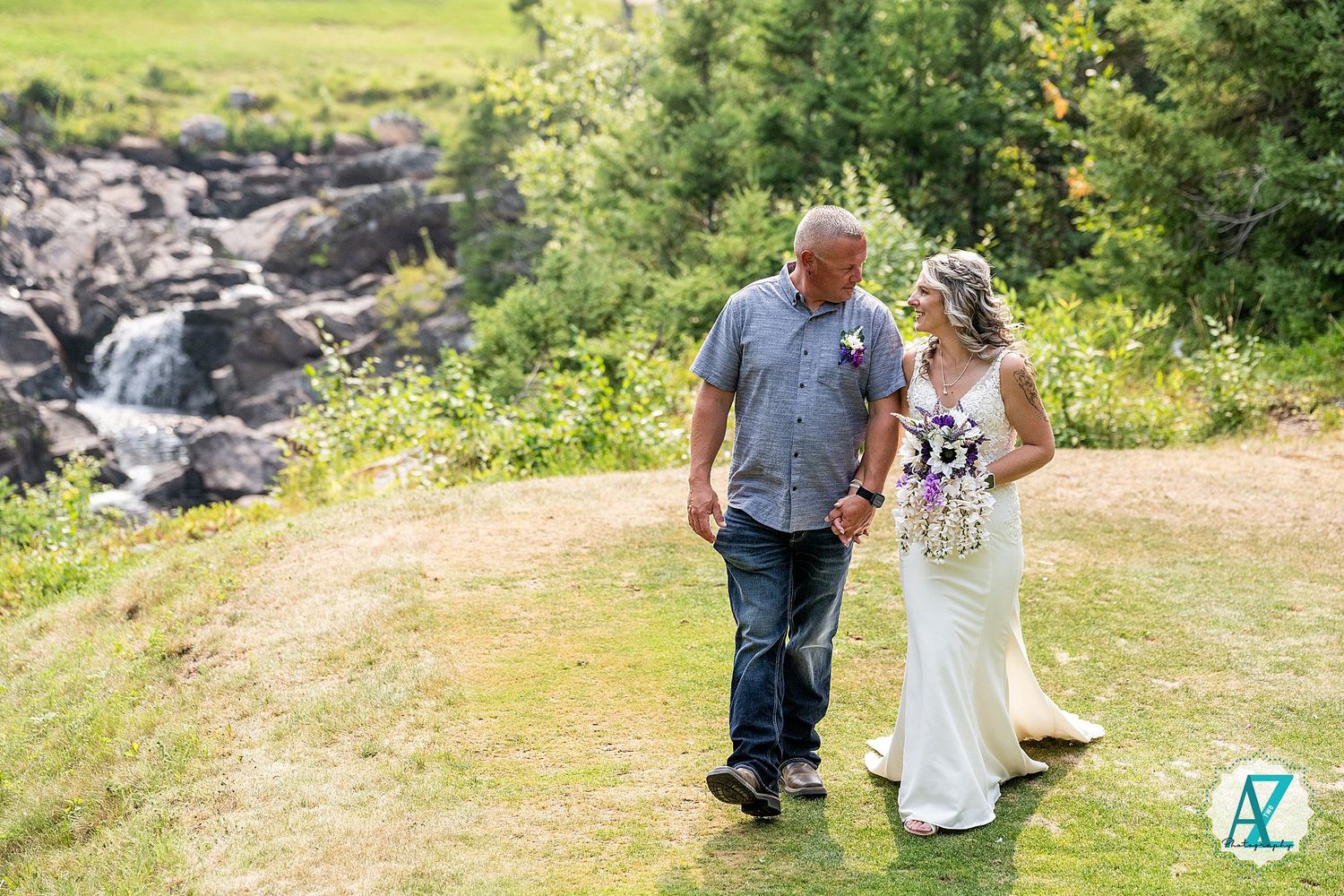 A couple walks along a grassy path near a rushing stream surrounded by lush greenery and rocks on a sunny day.
