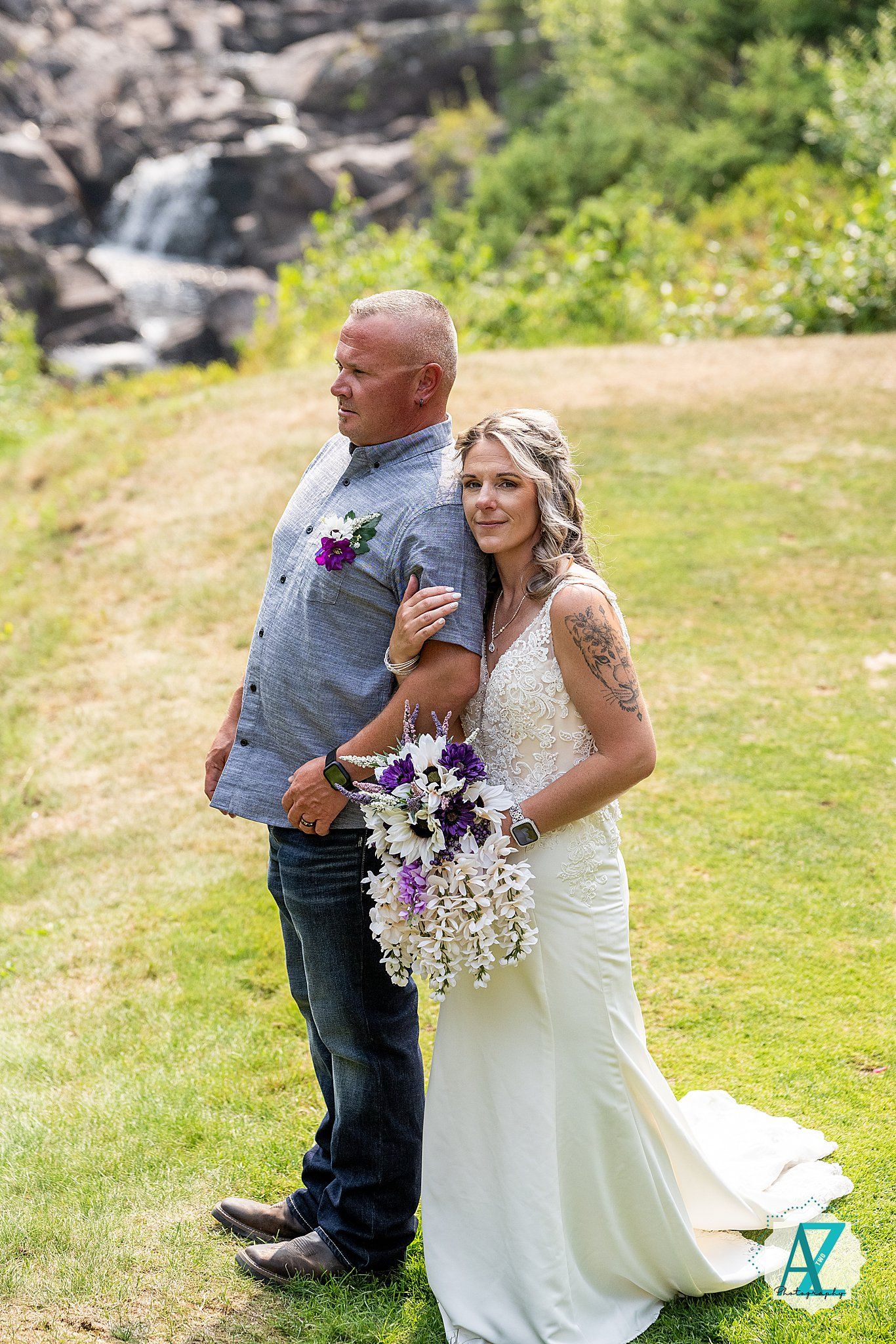 A couple shares a tender moment at their outdoor wedding ceremony near a waterfall, with purple floral bouquet accents.