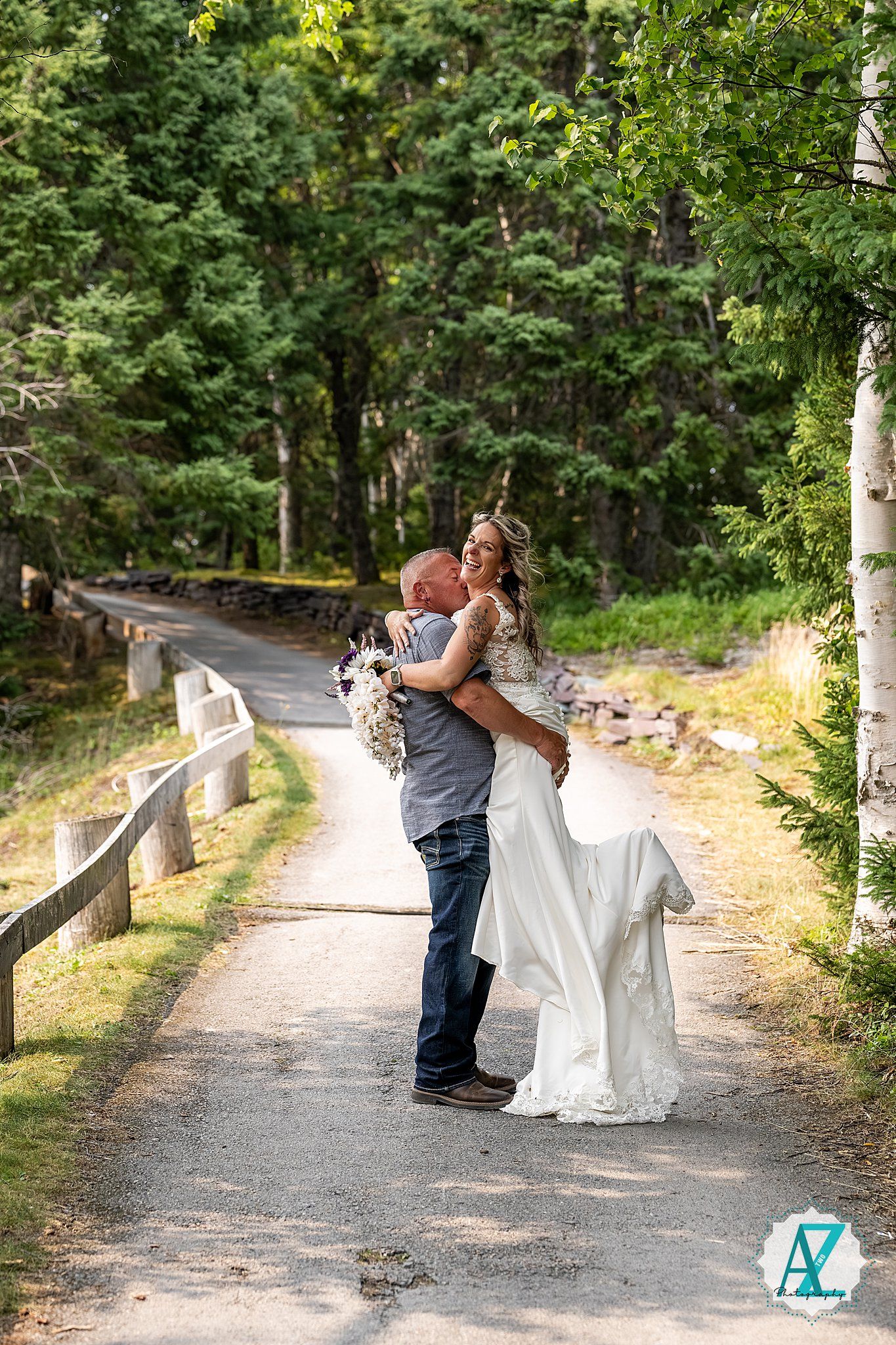 A couple embraces on a scenic woodland path surrounded by lush greenery and a rustic wooden fence on a sunny day.