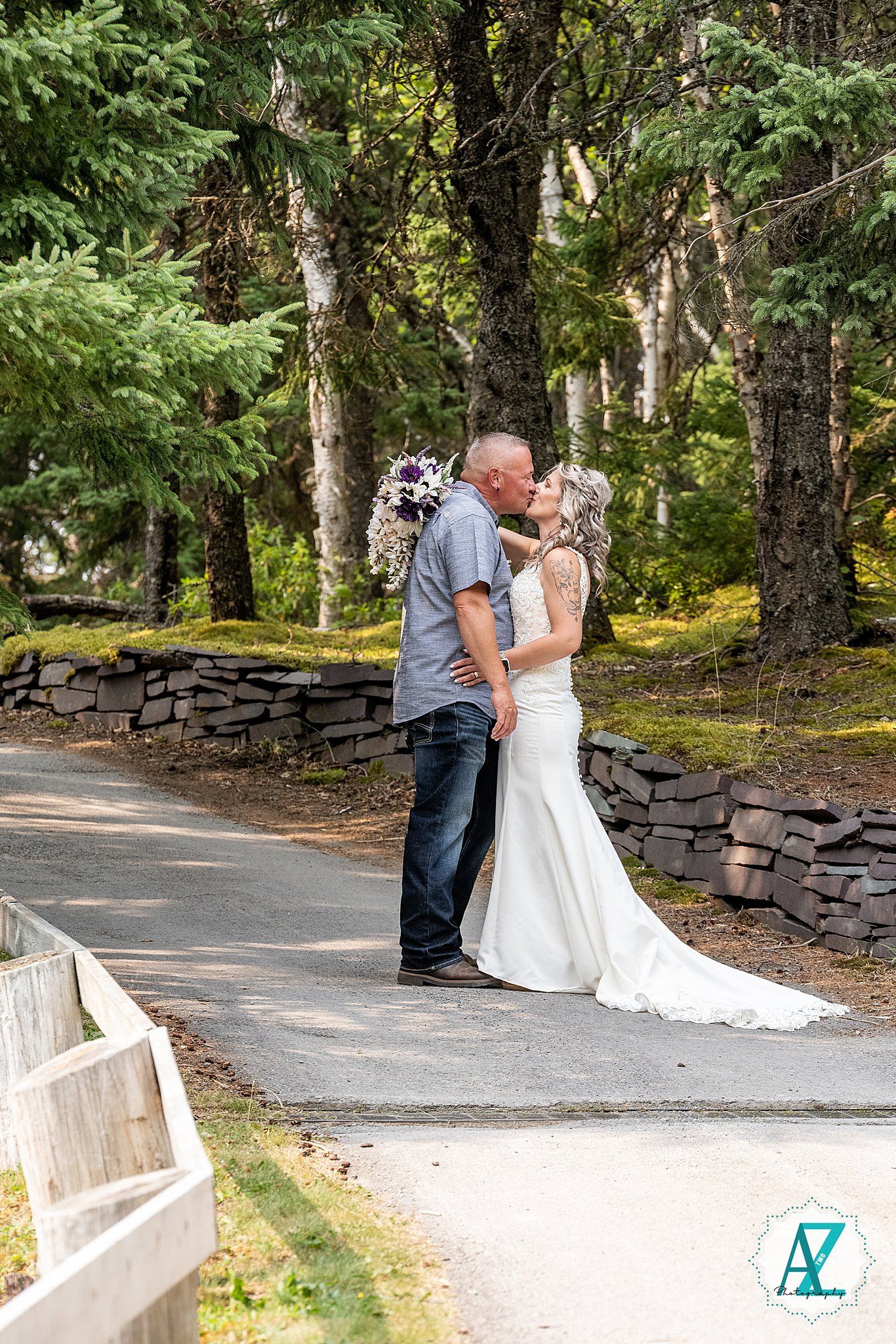A couple in wedding attire shares a romantic kiss along a stone-lined path in a wooded outdoor setting.