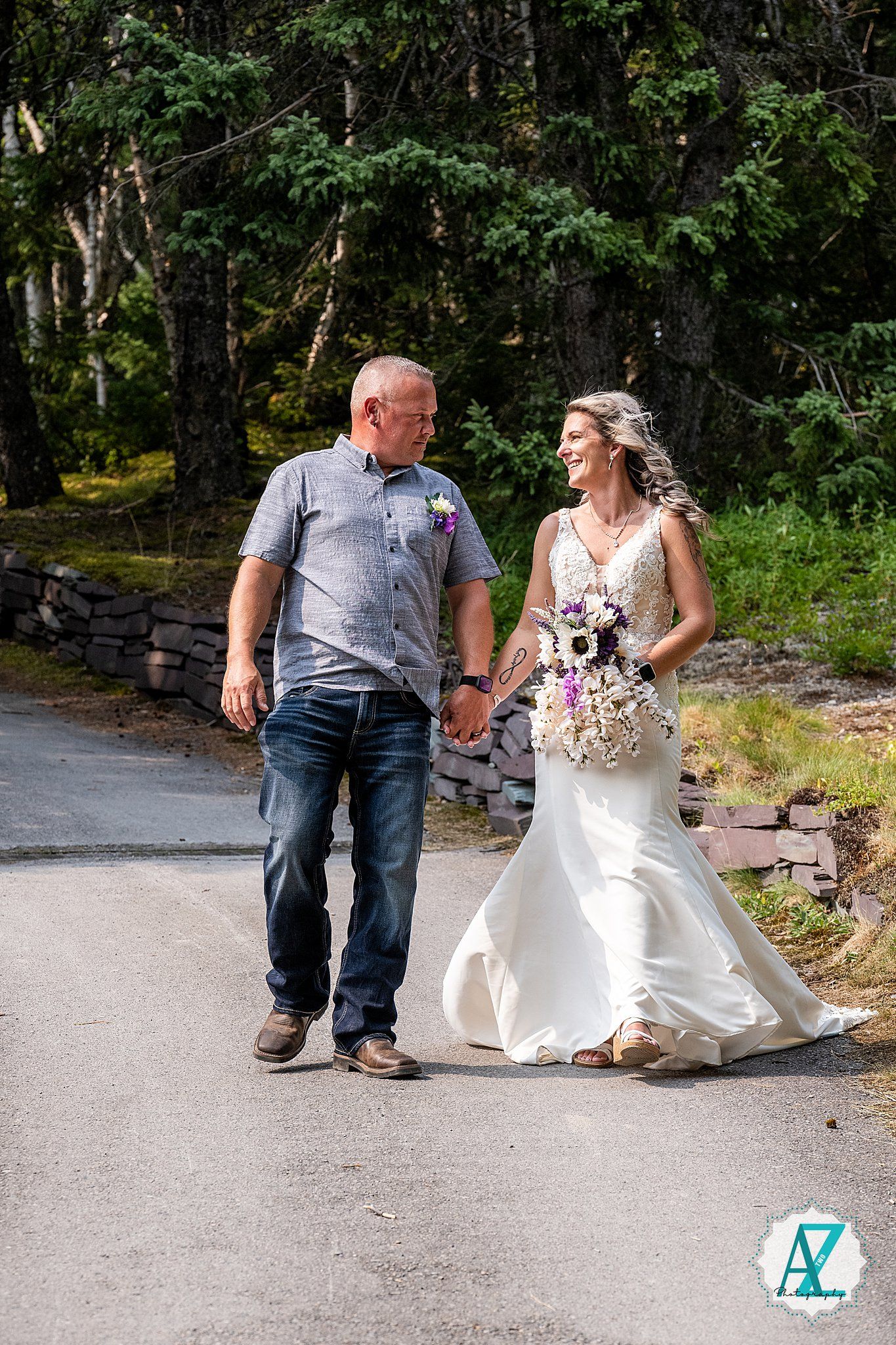 Two people walk down a paved path surrounded by green forest, one wearing a white wedding gown and the other in casual attire.