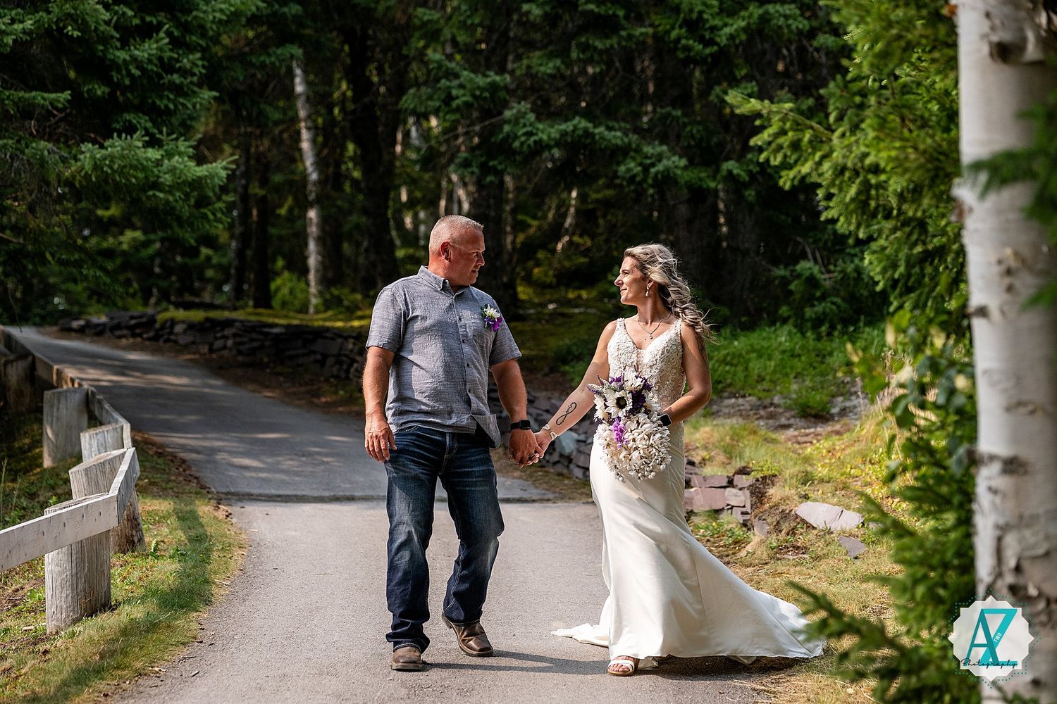 A couple walks hand in hand down a wooded path surrounded by birch trees and greenery on a sunny day.