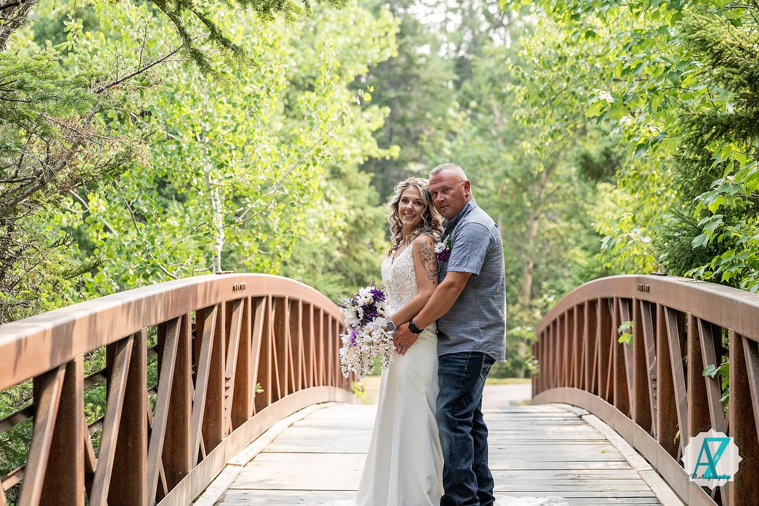 A couple embraces on a wooden bridge surrounded by lush green trees in a romantic outdoor setting.