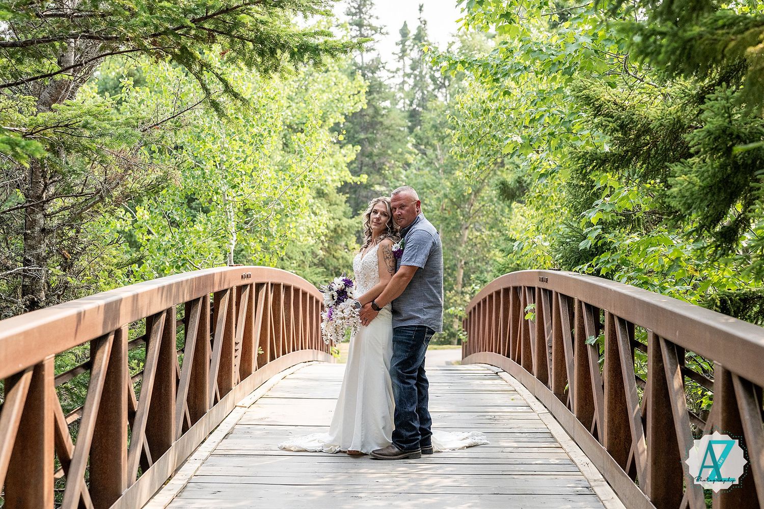 A couple shares a romantic moment on a wooden footbridge surrounded by lush green trees.
