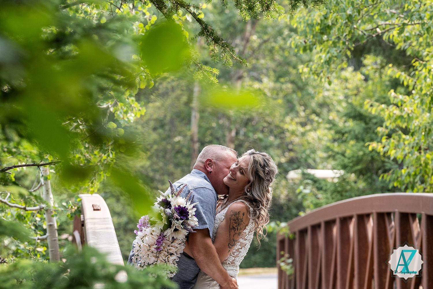 A couple shares a romantic kiss on a wooden bridge surrounded by lush green foliage on a sunny day.