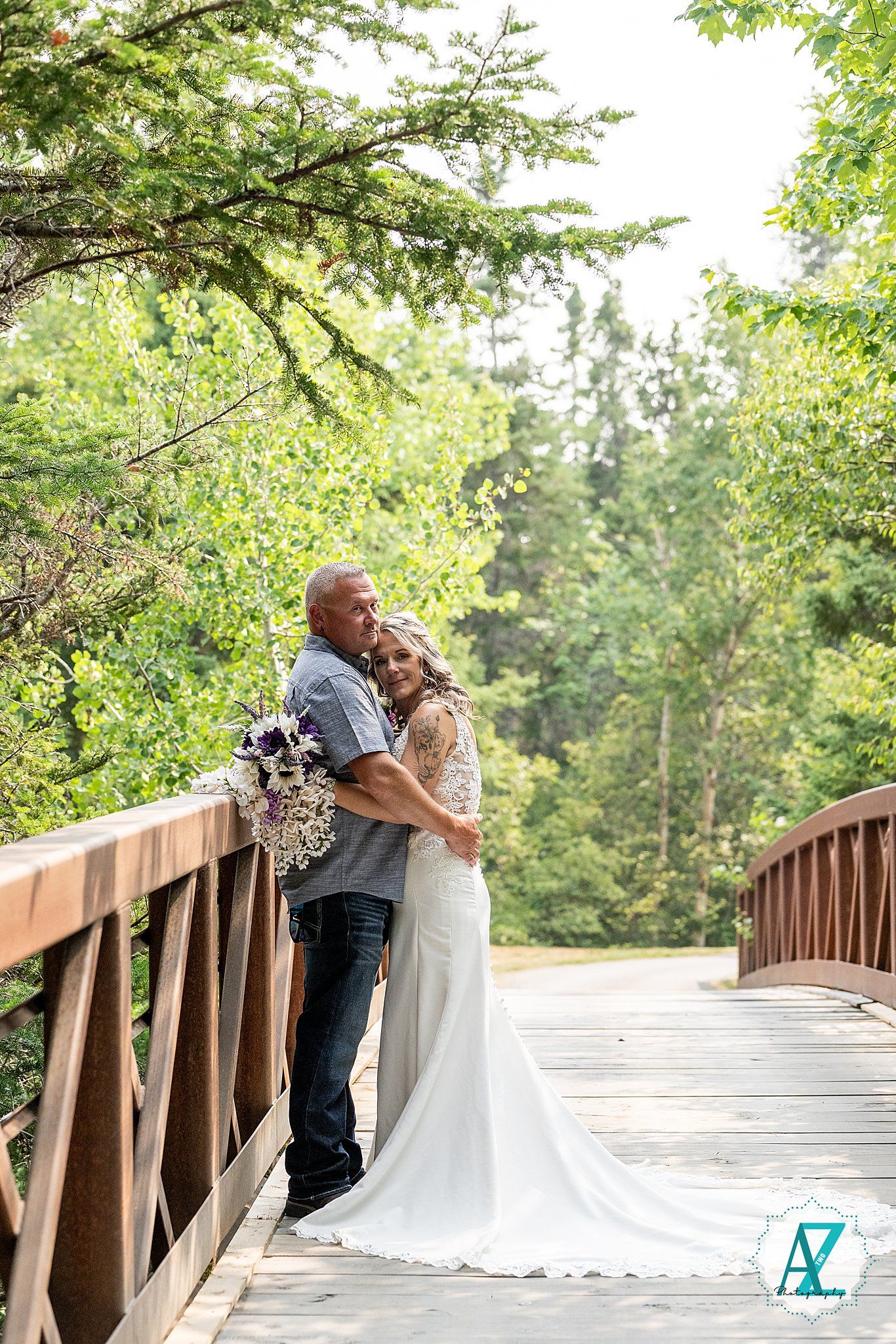 A couple embraces romantically on a wooden bridge surrounded by lush green trees on a bright sunny day.