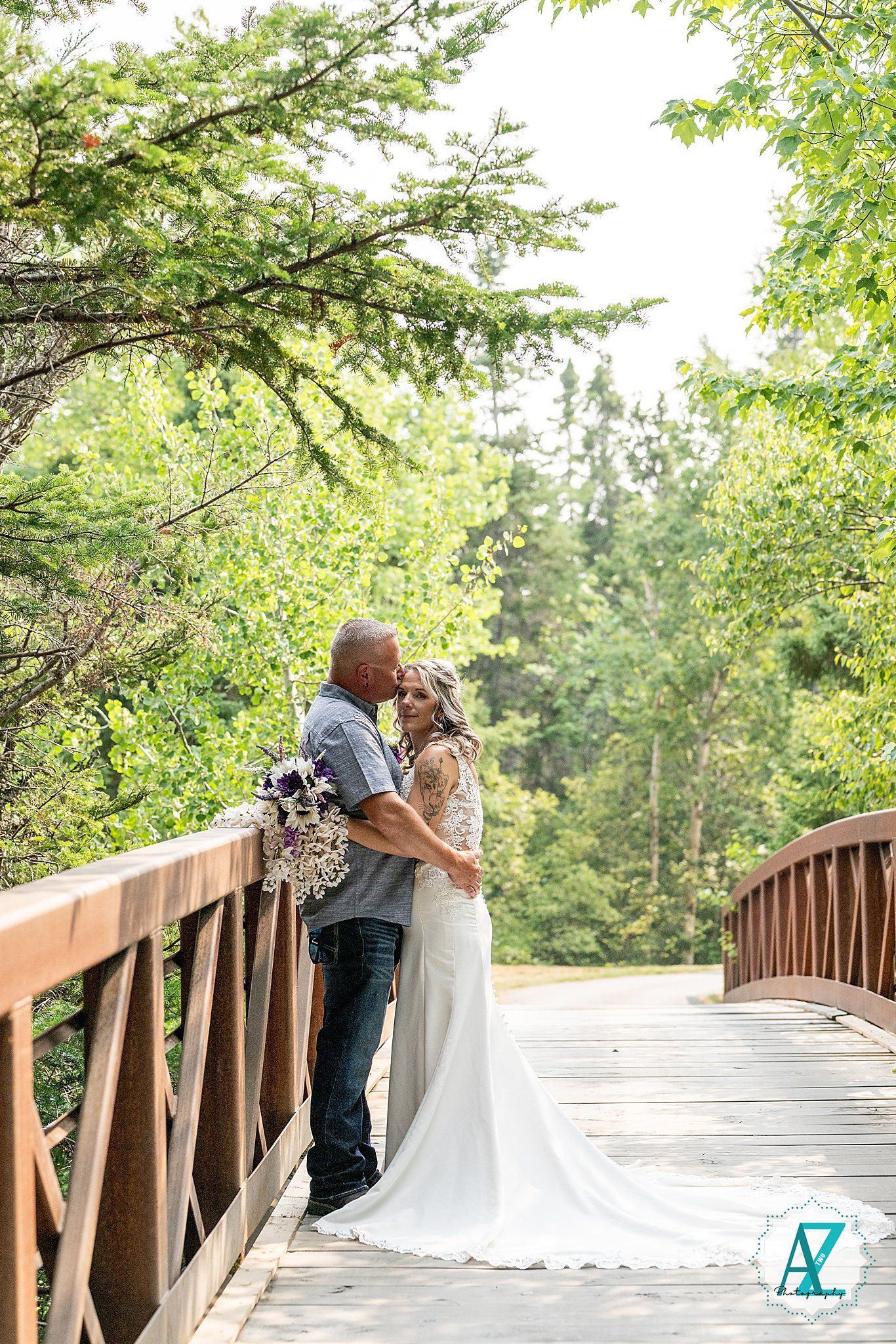 A couple shares a romantic moment on a wooden bridge surrounded by lush green trees in a natural outdoor setting.