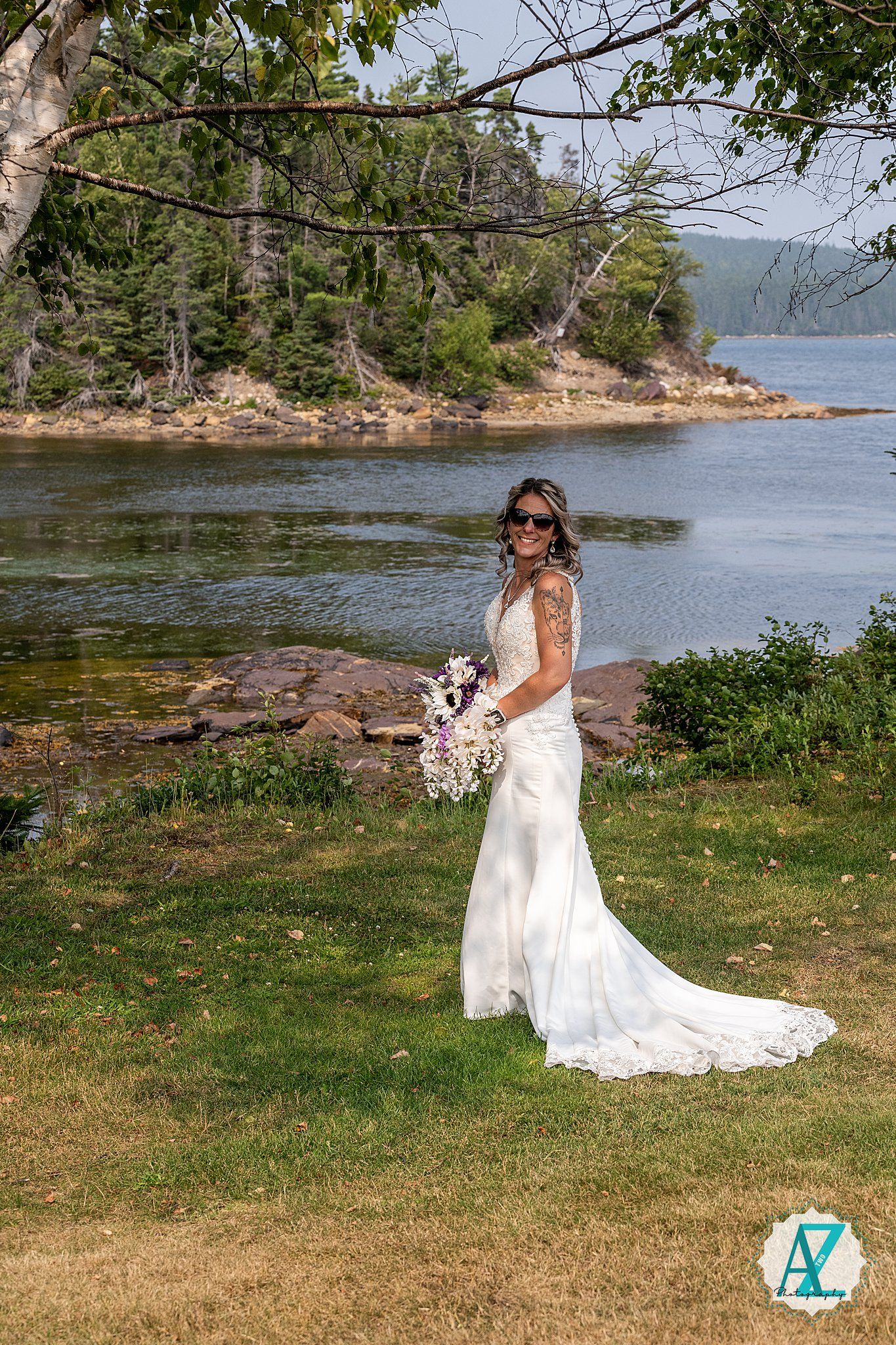A scenic coastal wedding photo showing an elegant white lace gown with a long train on a grassy shore by the water.