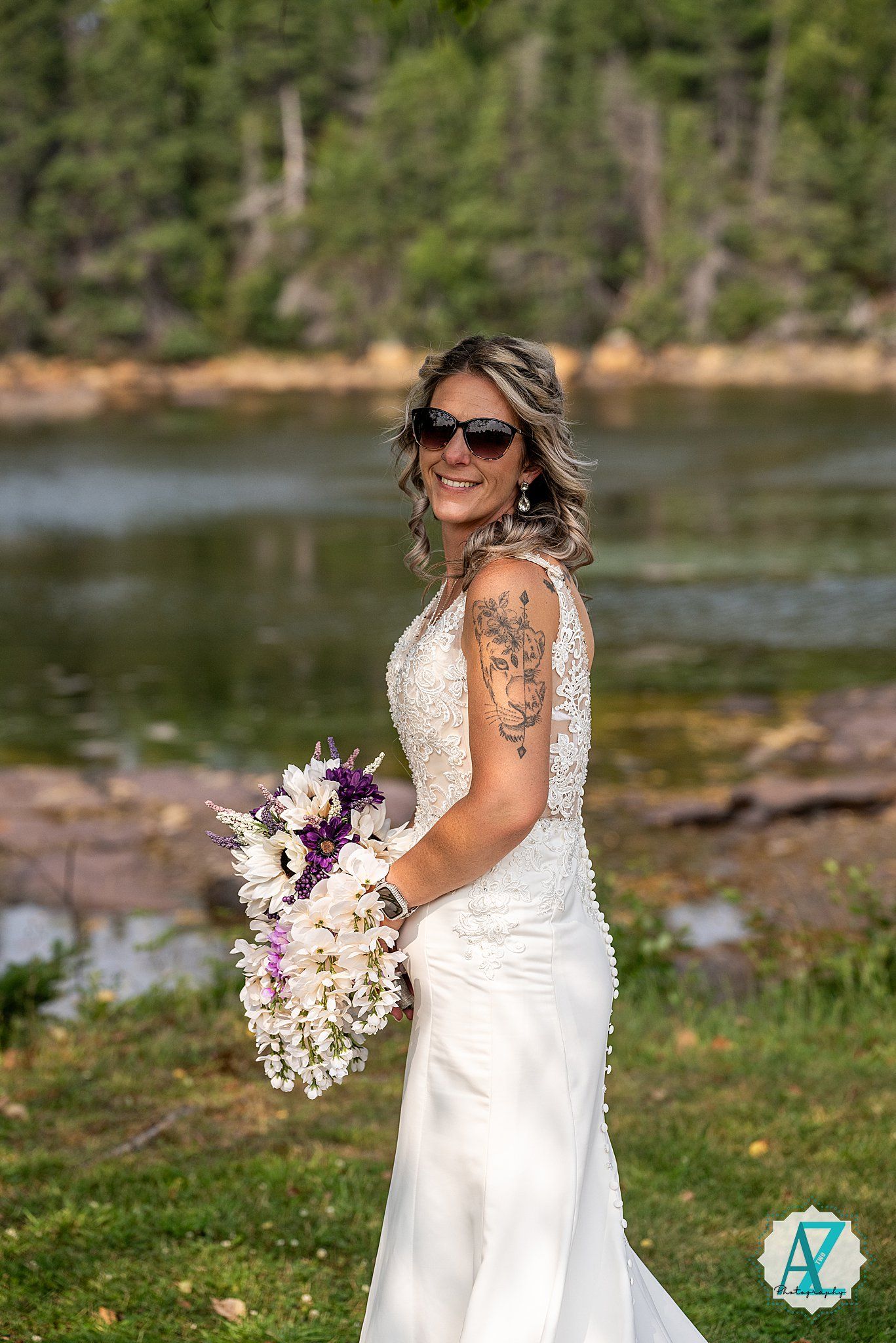 A bride in a fitted lace wedding gown and sunglasses holds a purple and white bouquet by a lakeside.