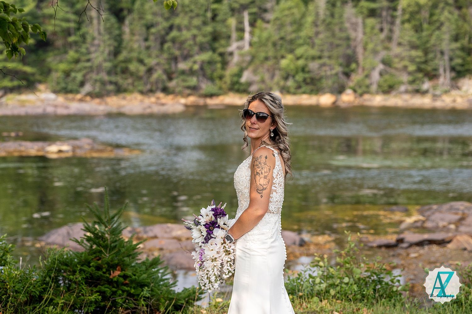A scenic wedding photo taken at a lakeside location surrounded by pine trees and rocky shores.