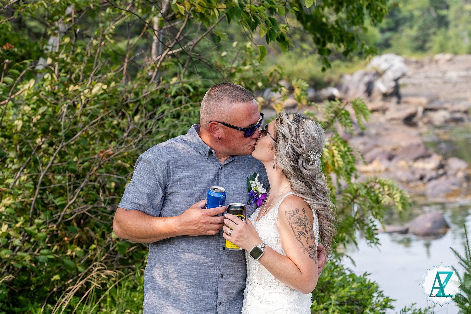 A couple shares a romantic moment with coffee cups by a rocky shoreline surrounded by green foliage.