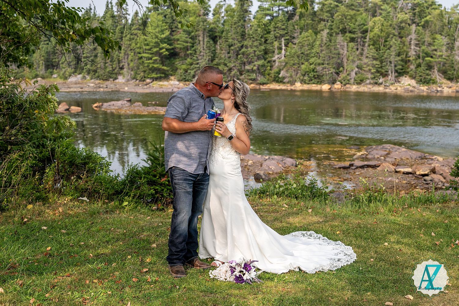 A couple enjoys an intimate moment by a serene lakeside in a wooded natural setting during their wedding day celebration.