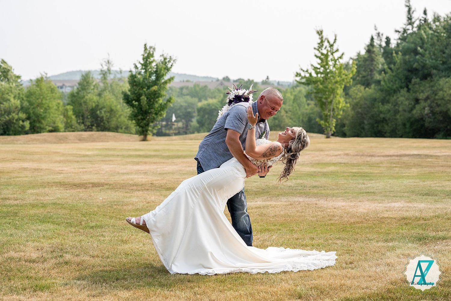 A couple shares a romantic dip pose on their wedding day in a scenic field with green trees in the background.