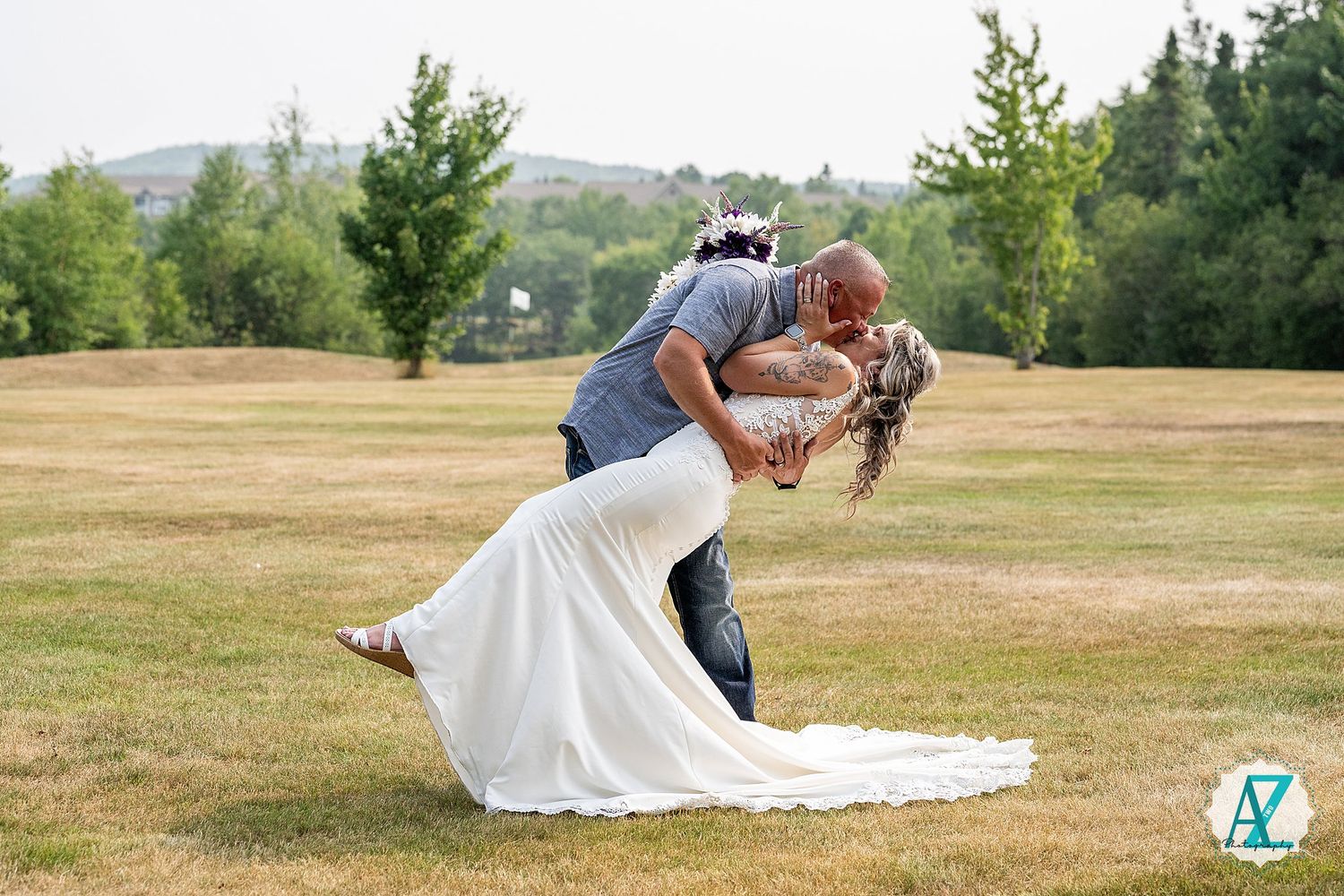 A couple shares a romantic dip kiss outdoors on a grassy hill surrounded by trees on a summer day.