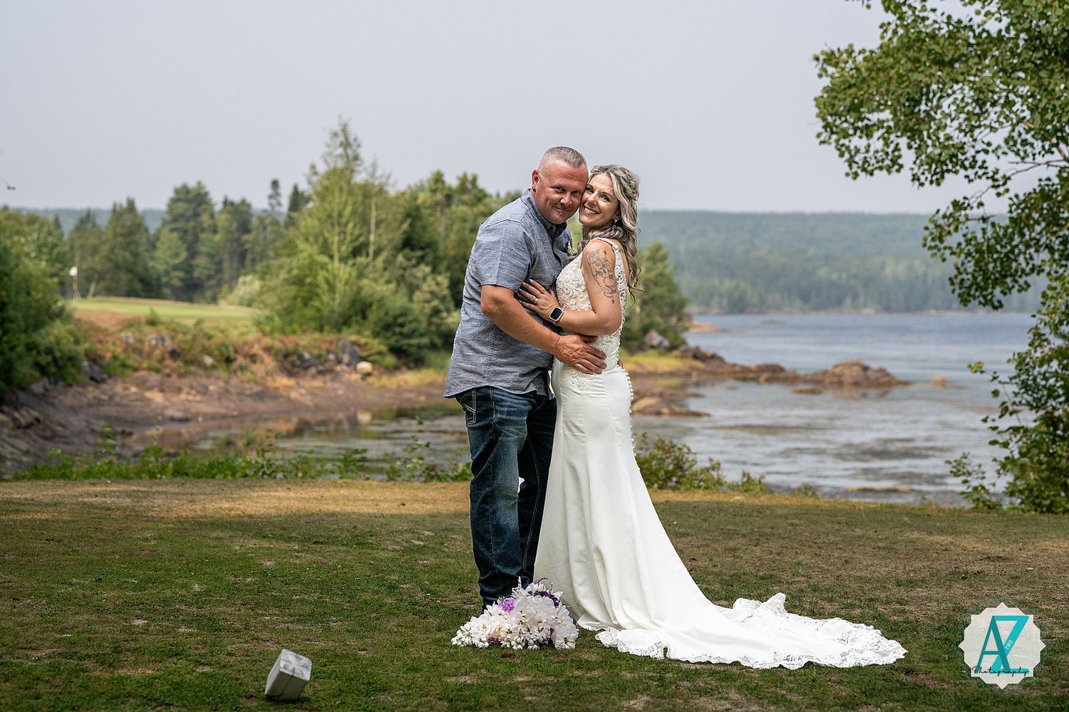 A couple embraces and kisses at a lakeside location with green trees and shoreline in the background.