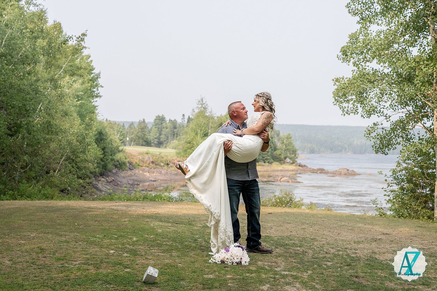 A joyful wedding couple embraces on a scenic grassy bluff overlooking a serene lake surrounded by trees.