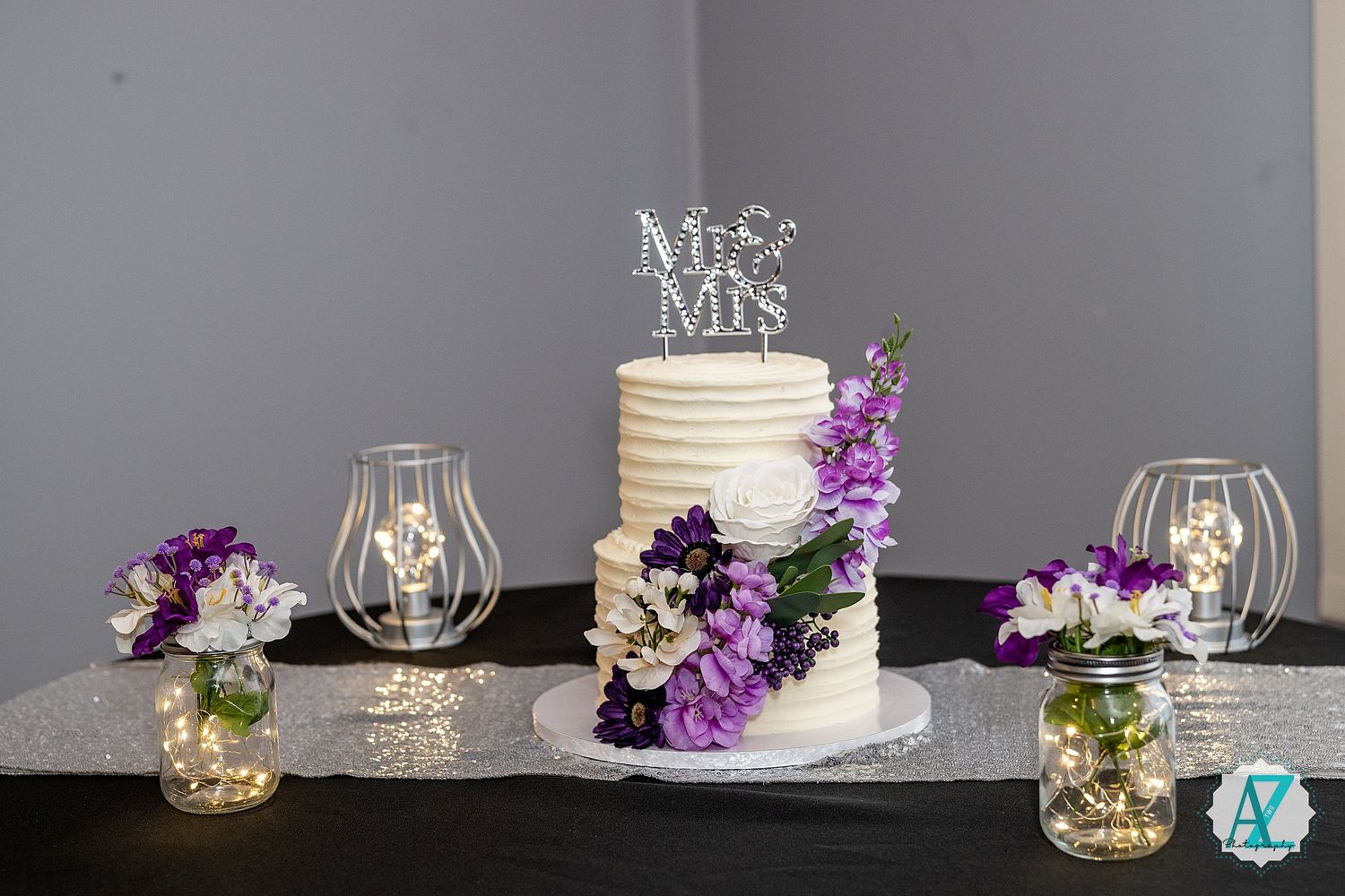 A two-tier white wedding cake decorated with purple and white flowers sits on a table with wine glasses and floral arrangements.