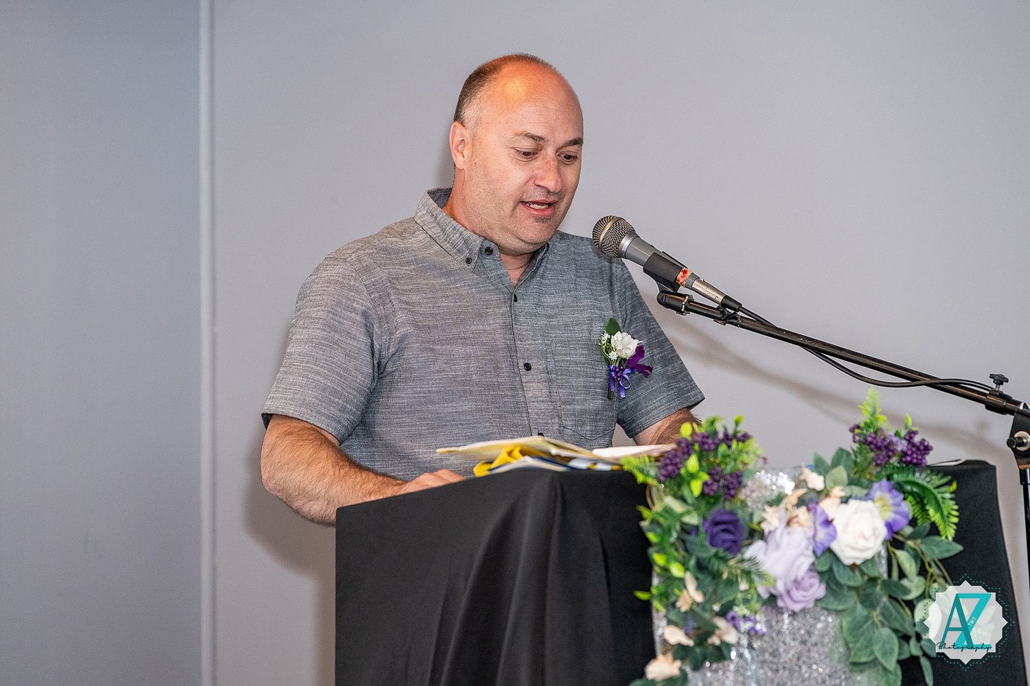 Speaker at podium reads from notes during event with floral decorations and microphone setup.