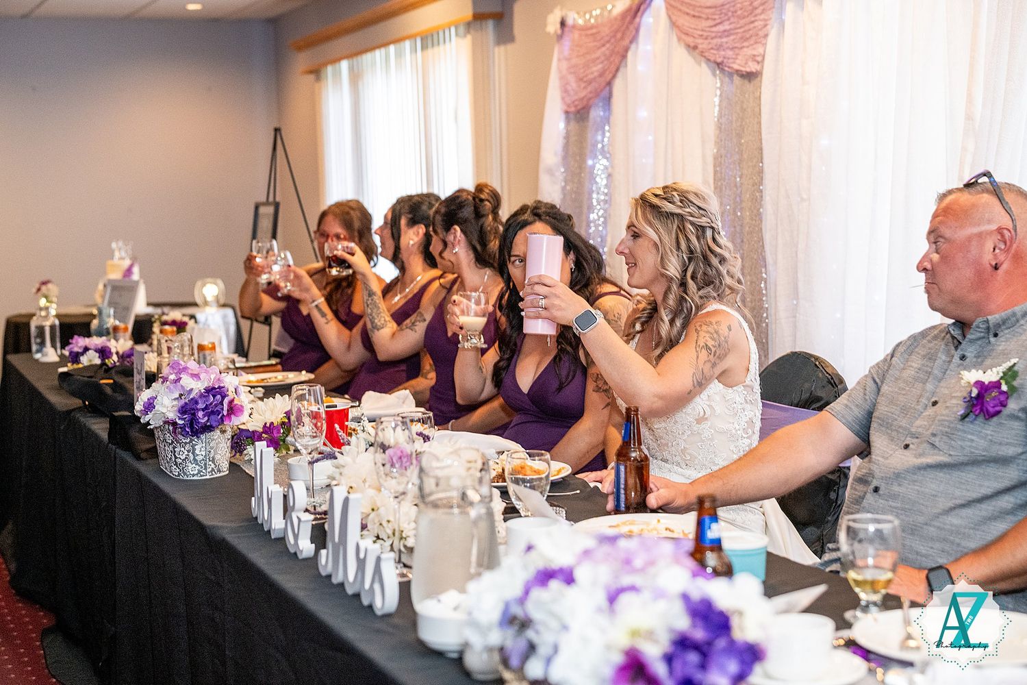 Wedding party members react with excitement while seated at a reception table decorated with purple floral centerpieces.
