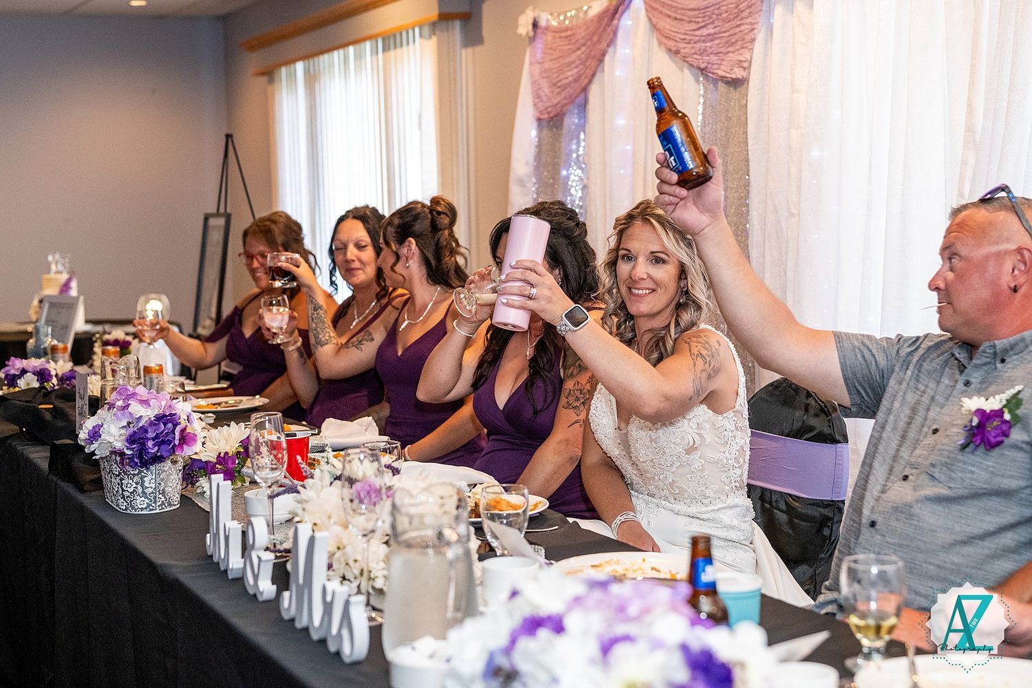 Wedding party members gathered at a reception banquet table with black tablecloth and purple floral decorations.