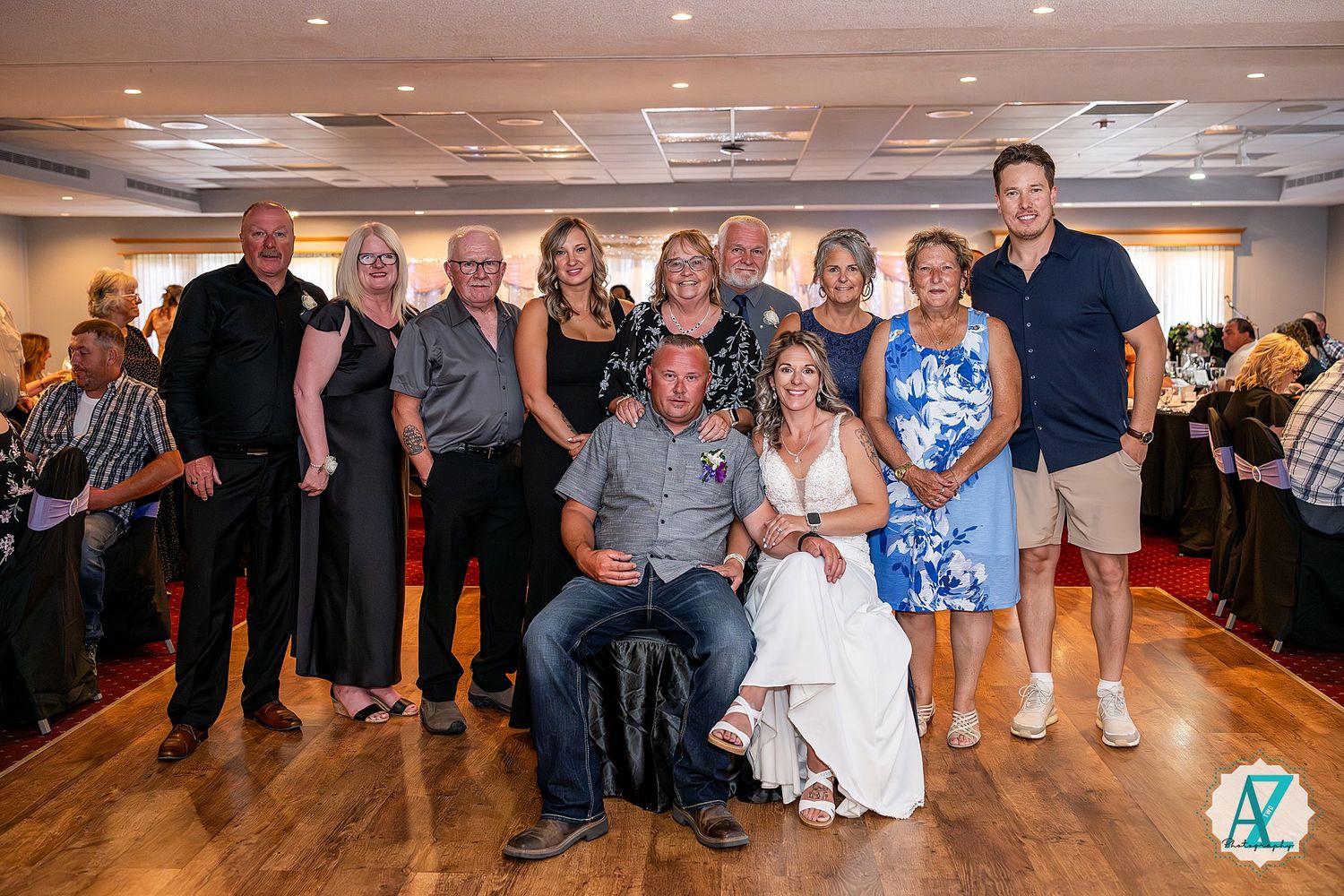 A group poses together for a formal celebration photo on a wooden dance floor at an indoor event venue.