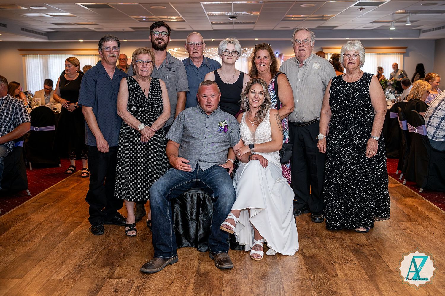 A group photo at a wedding reception with people gathered on a wooden dance floor in formal attire.