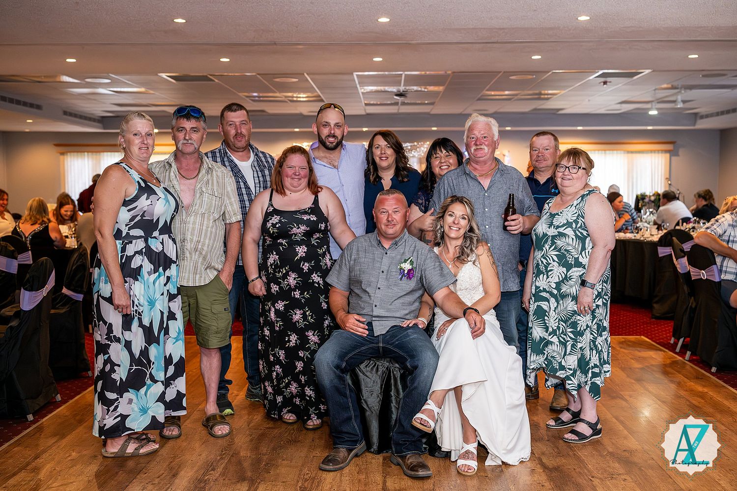 A group of wedding guests wearing coordinated blue floral dresses and casual attire pose together on a wooden dance floor.