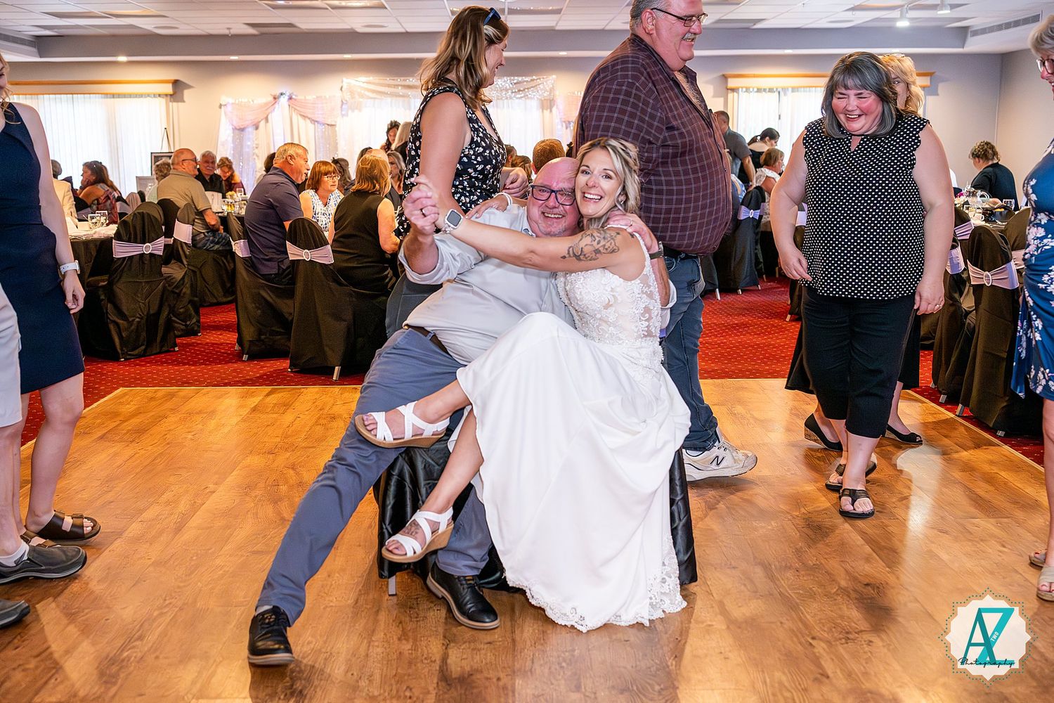 Wedding guests dance and celebrate on a wooden dance floor during a reception in an indoor venue with a red carpet.