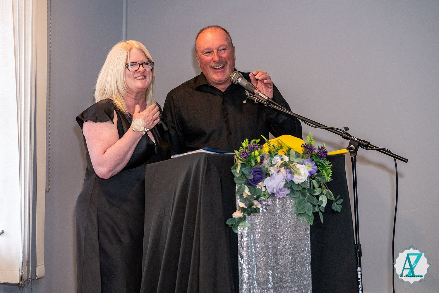 A group performs at an event with microphones on stage, decorated with floral arrangements and silver decor.