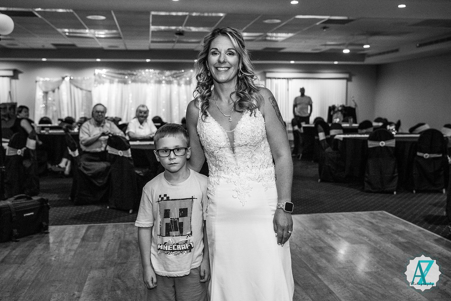 A bride in a white lace dress stands with a young boy wearing glasses in a reception hall with other guests visible.