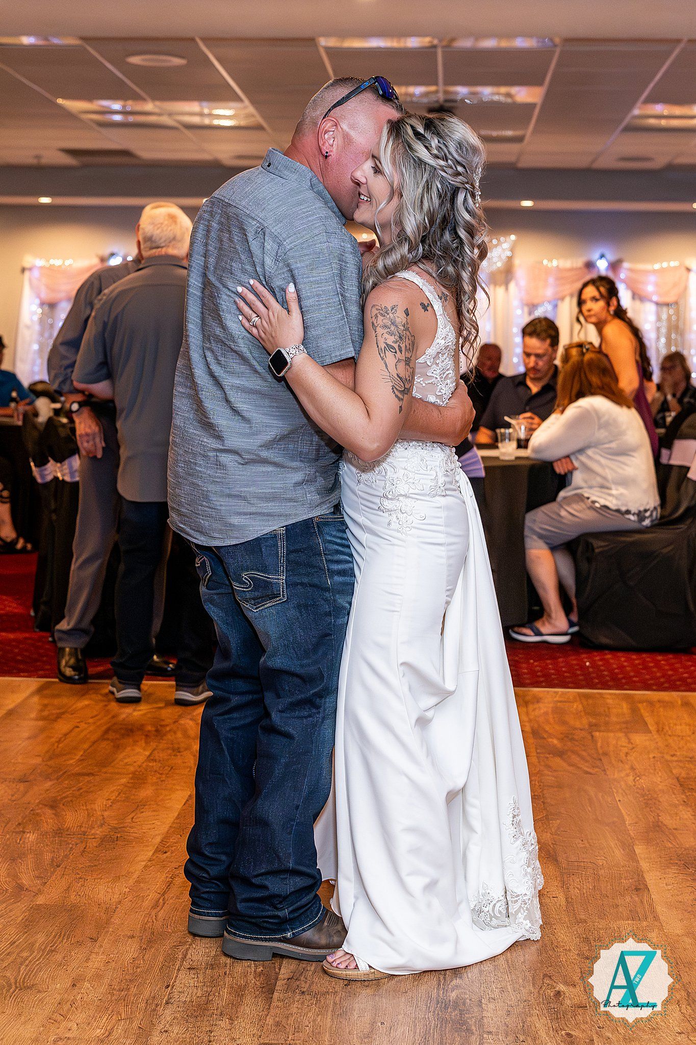 A couple shares their first dance at a wedding reception on a wooden dance floor with guests in the background.