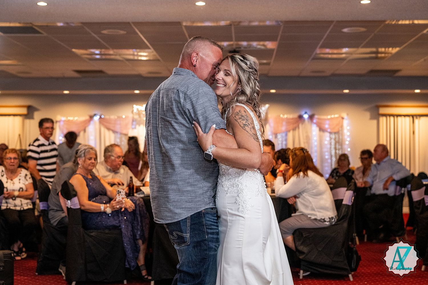 A couple shares a tender dance moment in a wedding reception banquet hall filled with guests.