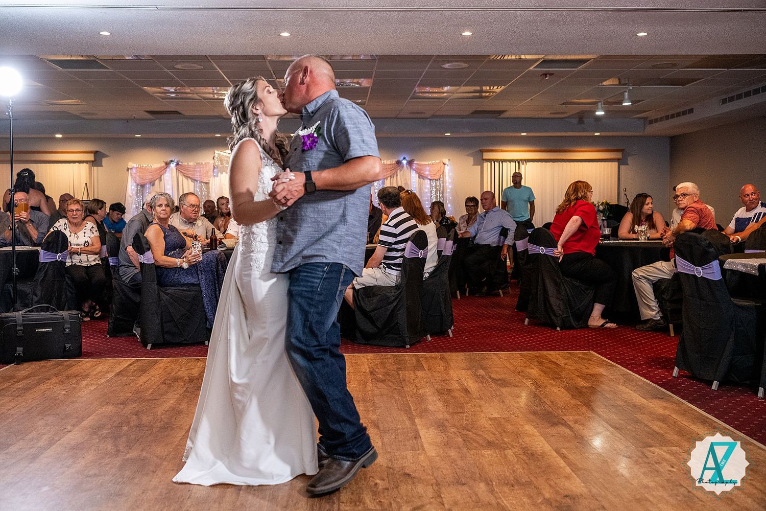A wedding party performs synchronized group dance moves on a wooden dance floor in a banquet hall.