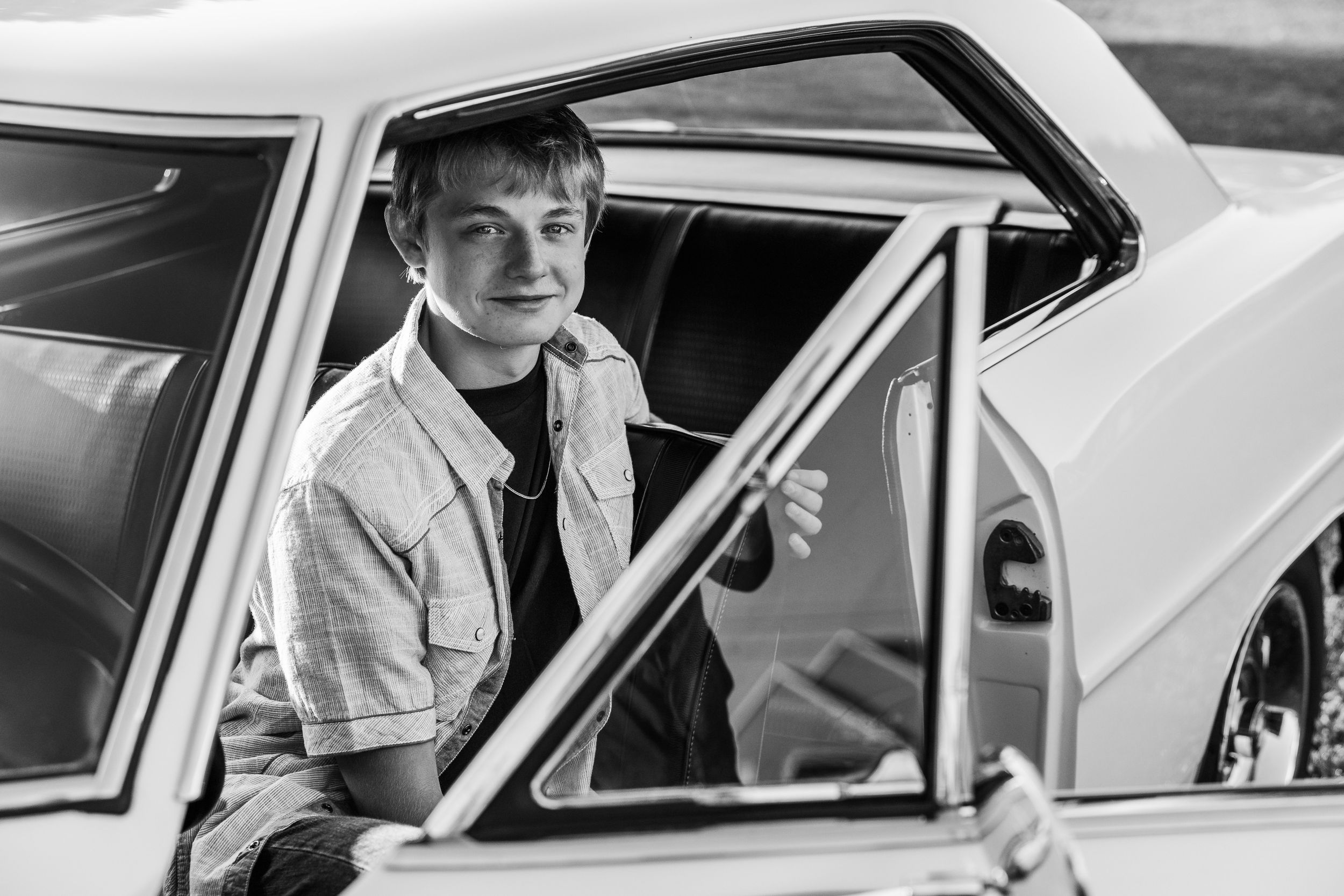 A vintage black and white photo showing someone sitting in a classic car with the door open.