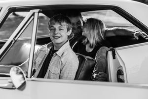 Two young people sit in a vintage car together laughing and smiling in black and white photography.