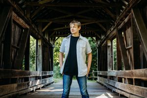 Someone standing in the middle of a wooden covered bridge wearing a black shirt, grey button-up, and jeans.
