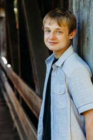 Young person in light blue button-down shirt smiles while leaning against wooden wall in outdoor setting.