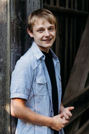 Young person in light blue button-up shirt over black t-shirt smiling while standing against dark background.