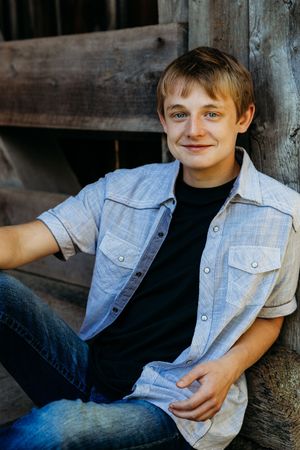 Young person in light denim button-up shirt and jeans sits casually against wooden wall in outdoor portrait.