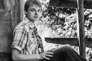 A young boy in a plaid shirt looks into the distance while sitting outside in a rustic wooden setting.