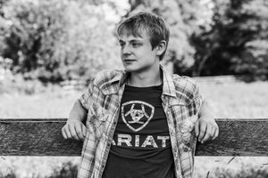 A young person in a plaid shirt and Ariat t-shirt leans against a wooden fence in a black and white photograph.