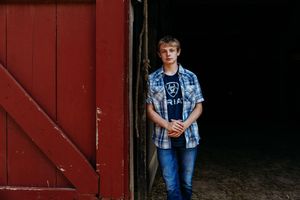A group of children pose in an outdoor barn setting wearing casual blue clothing against a red rustic backdrop.