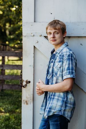 A young person in a blue plaid shirt and denim poses against a white porch column in the sunlight.