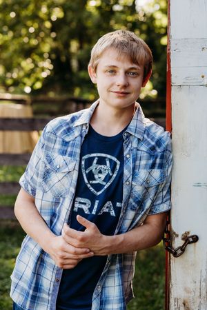 Youth in blue plaid shirt and navy t-shirt leans against white post in outdoor setting with bokeh background.