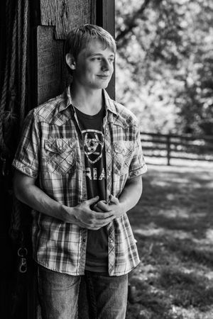 Black and white portrait of a person in casual plaid shirt leaning against wooden post, looking off to the side in natural setting.