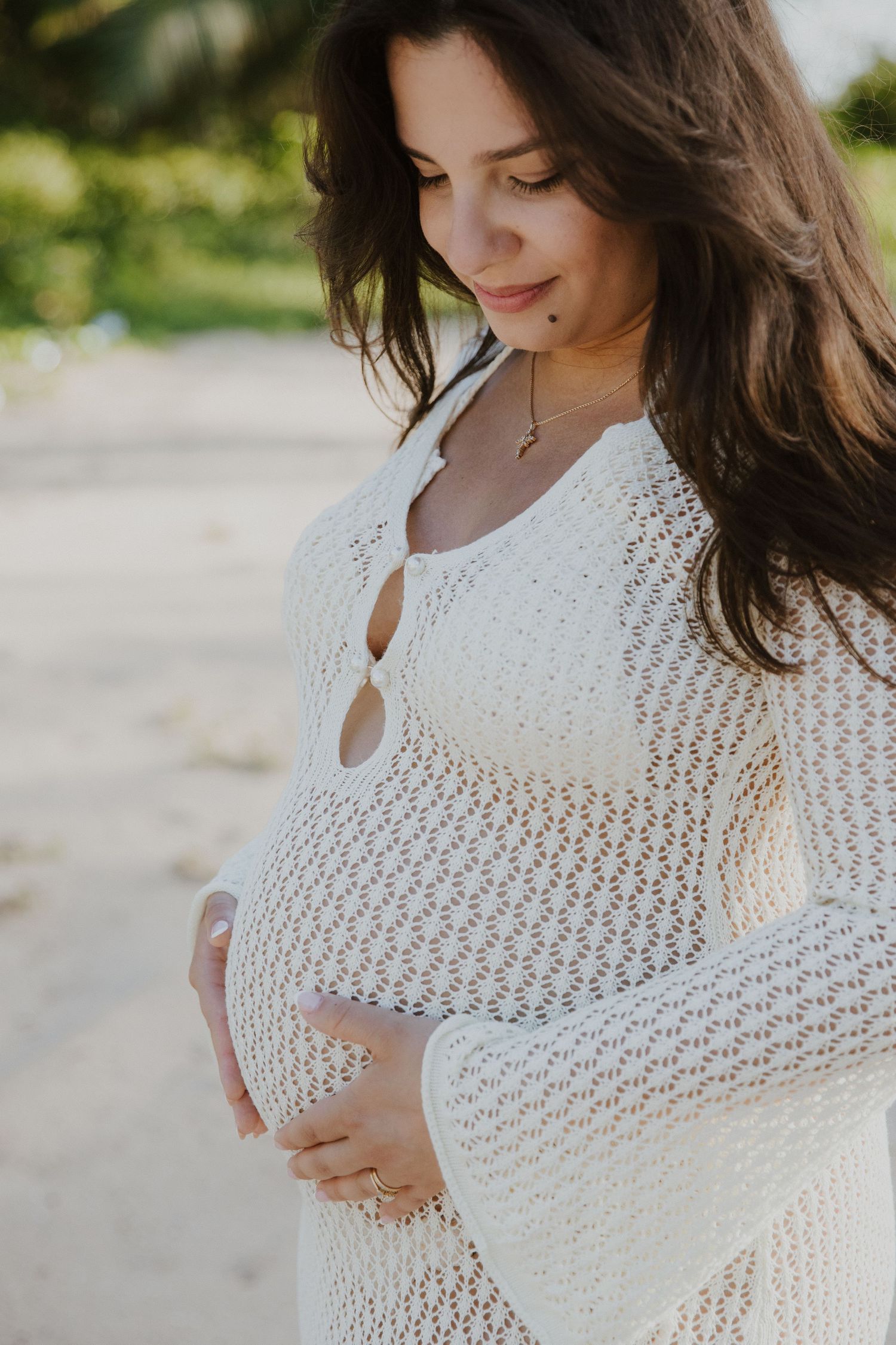 A maternity portrait in a white crochet dress on a sunny beach.