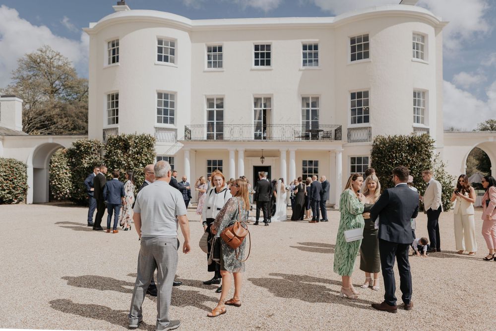 Wedding guests mingling outside a stunning white Georgian manor house on a sunny day.