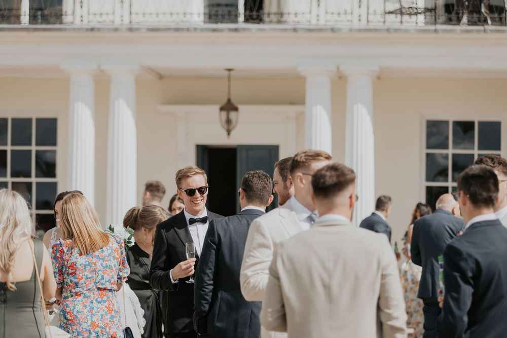 Elegantly dressed wedding guests socializing on the steps of a white columned manor house.
