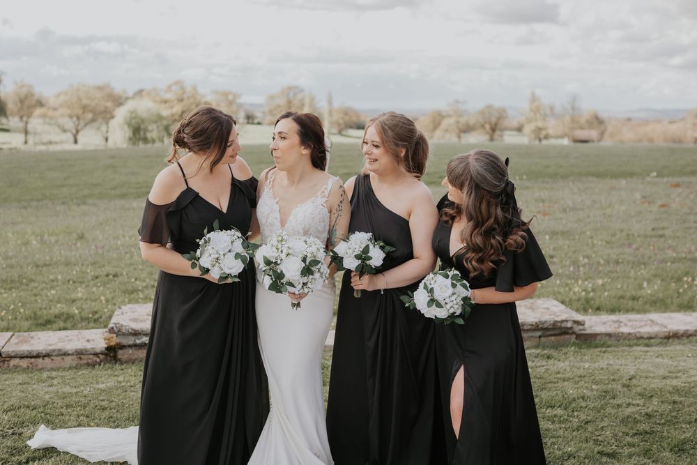 Bride with three bridesmaids in black dresses holding white bouquets in an open green field.