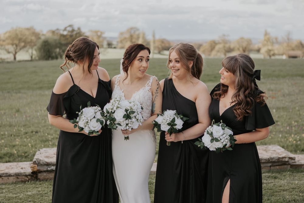 Bride and three bridesmaids in black gowns laughing together outdoors on a cloudy day.